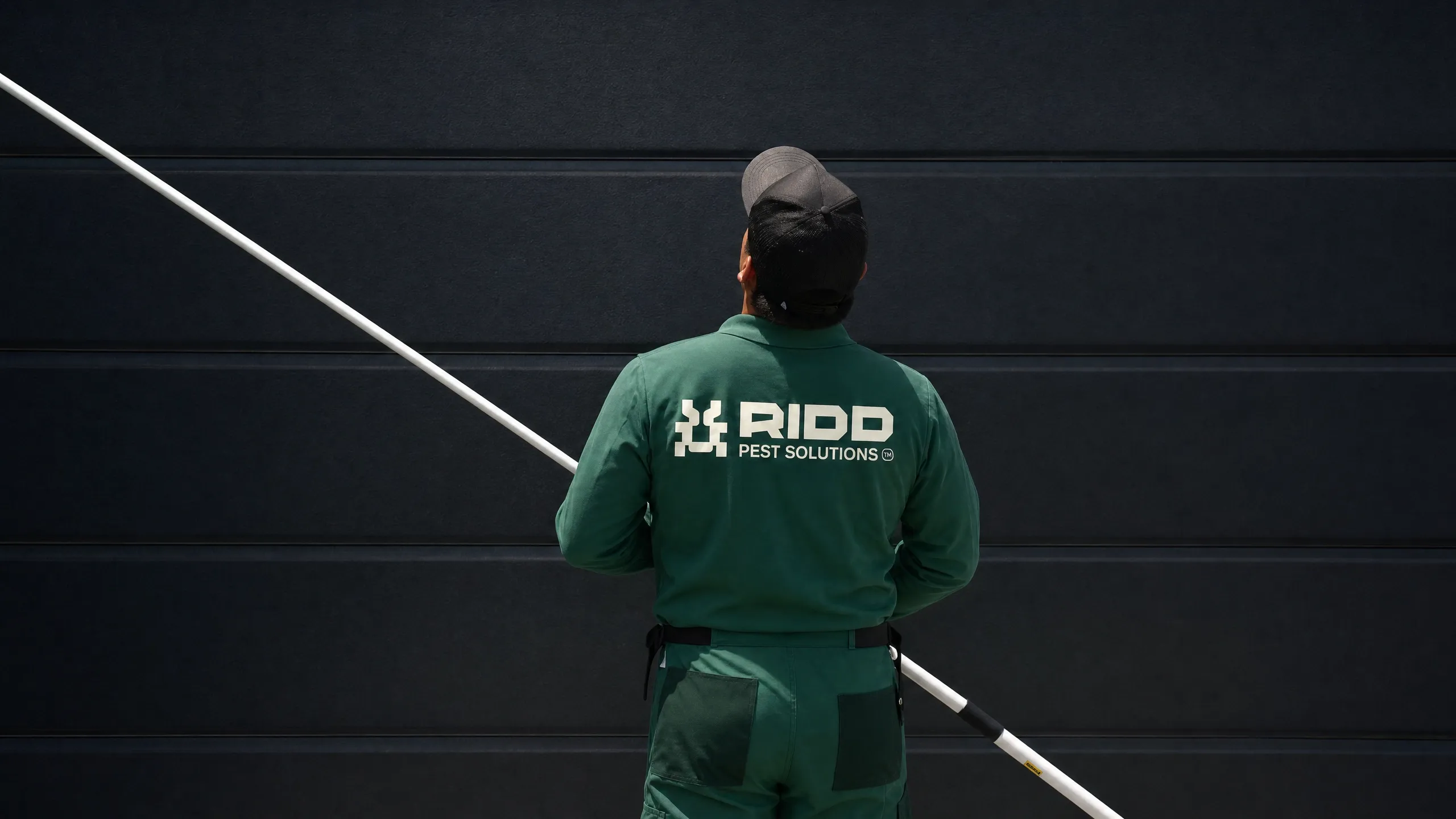 A RIDD pest control technician in a branded green uniform using an extension pole to treat the exterior of a home.